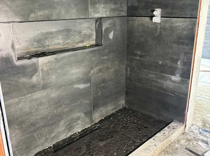 Bathroom shower mid-renovation with dark tile walls and pebble-tile flooring being installed